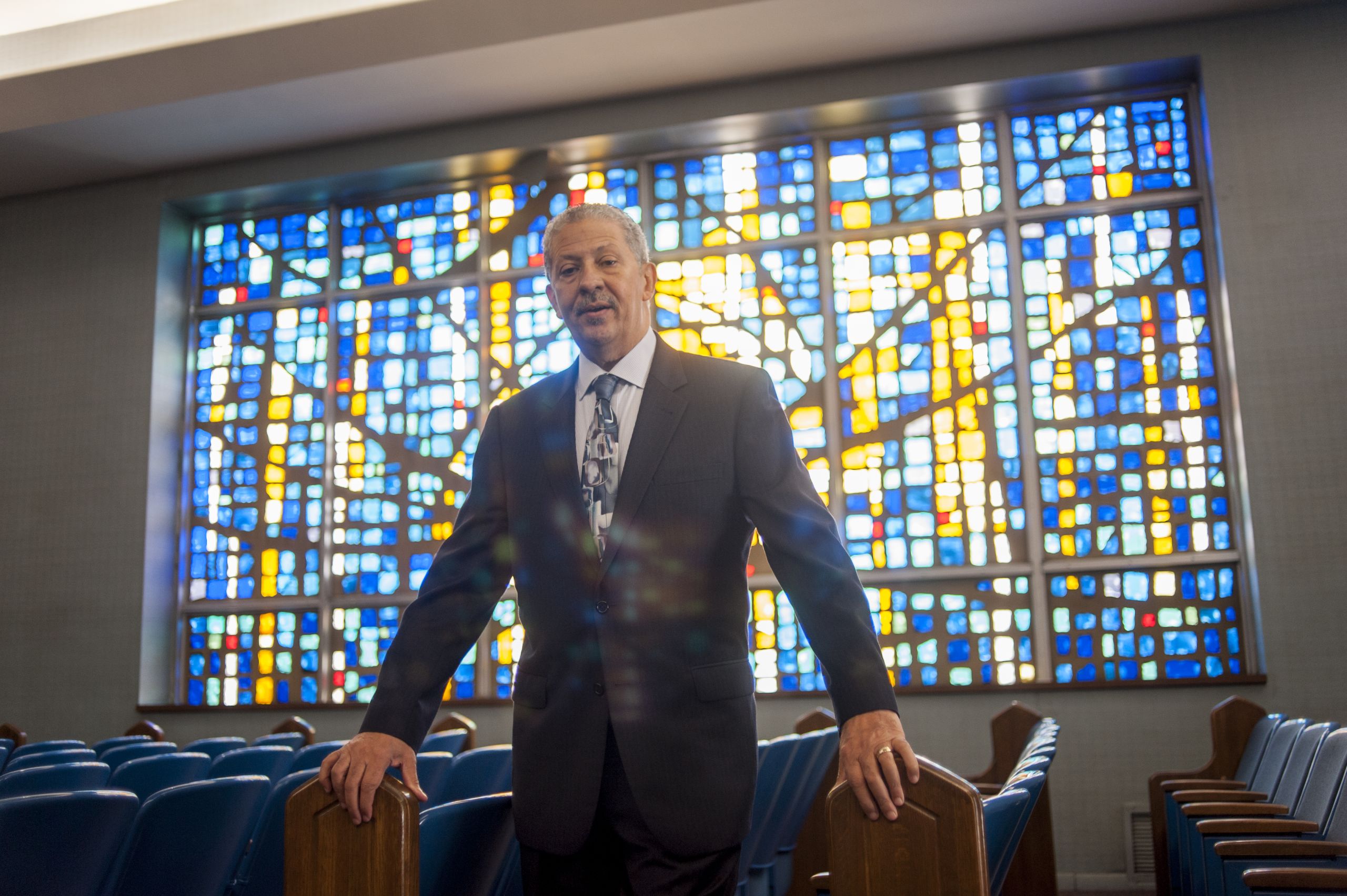 Norman Williams wears a suit and tie, looking directly into the camera. Behind him, a blue and yellow stained-glass window glows brightly.