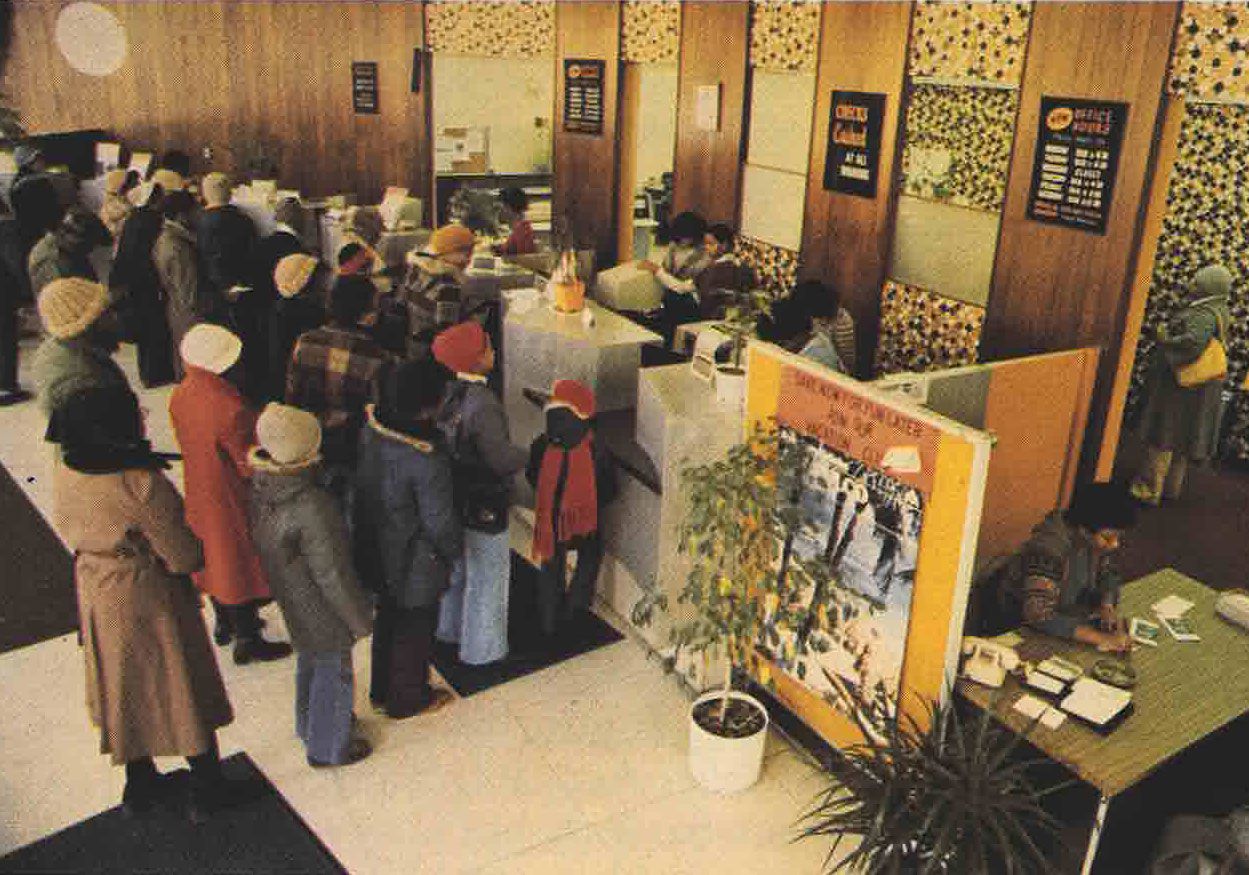 Customers line up at the counters in the Illinois Federal lobby. Many wear hats and coats. Plants and wood paneling decorate the lobby.