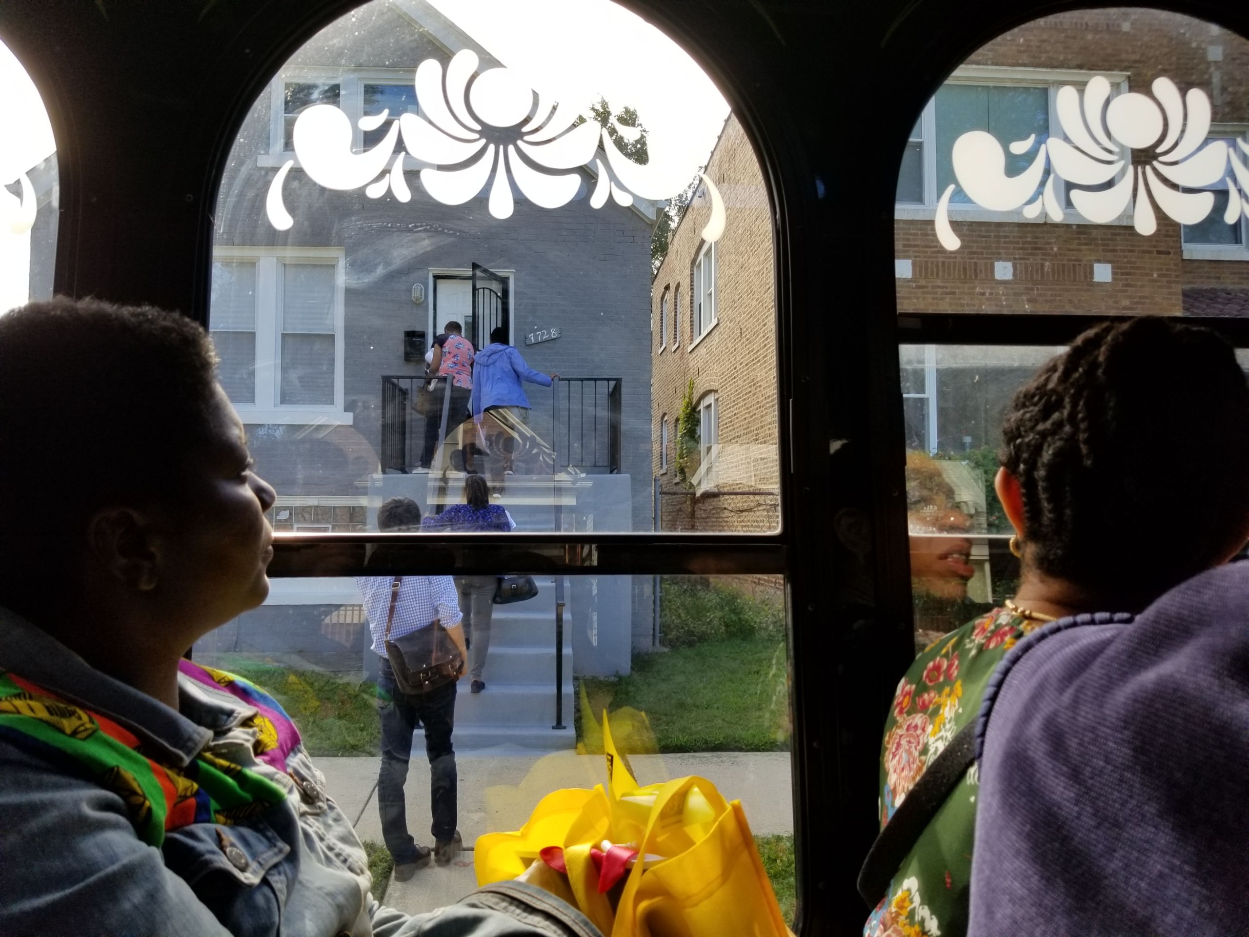 Through a the window of a trolley, the image shows people filing into a home in Chatham to look inside. Inside the trolley, others look at the house out of teh window.