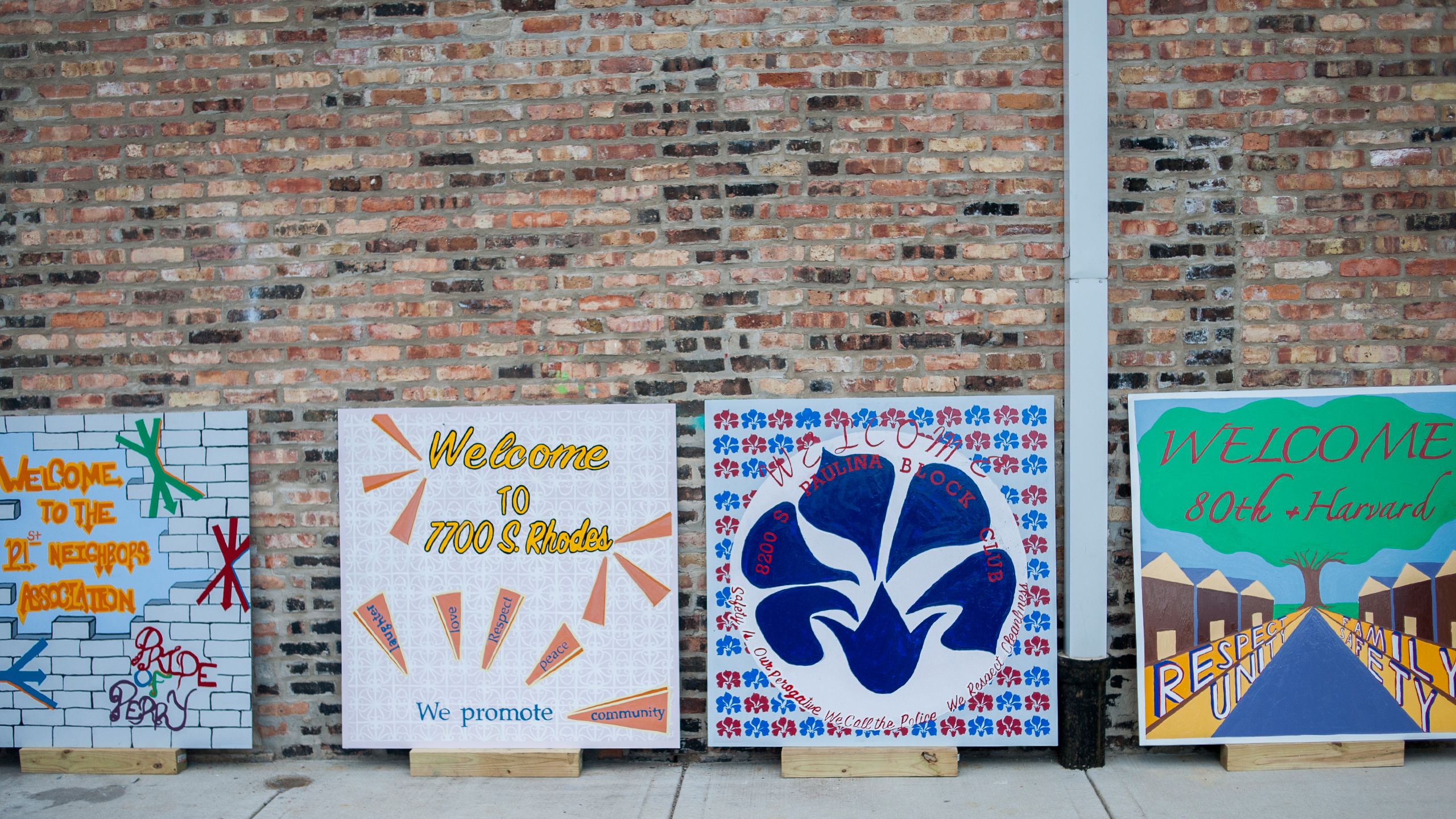 Colorful square signs with messages like "Welcome to 7700 S. Rhodes" lean up against a brick wall