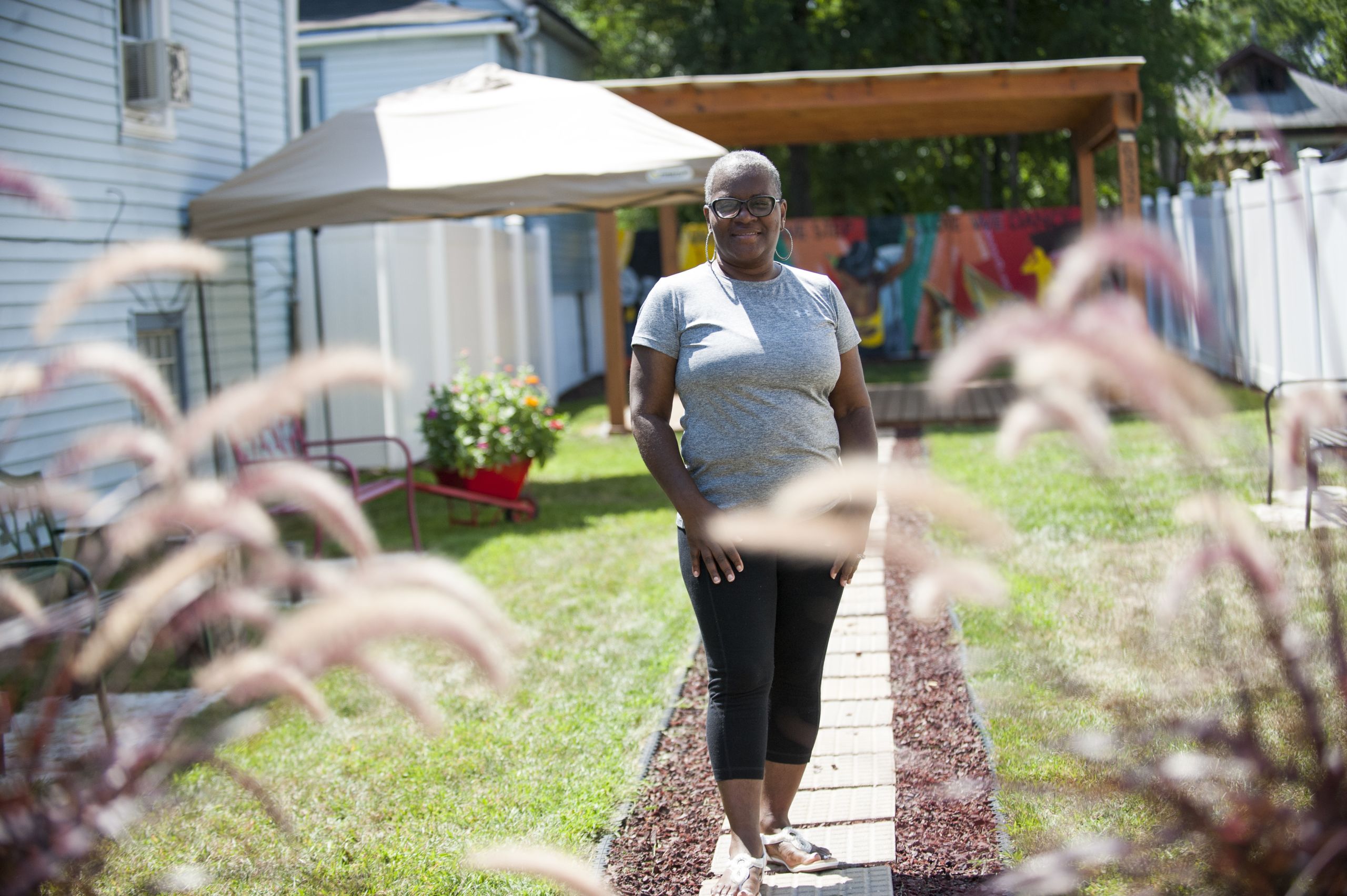 Tina Hammond, wearing a gray shirt, stands in her garden. There are pink plants in the foreground.