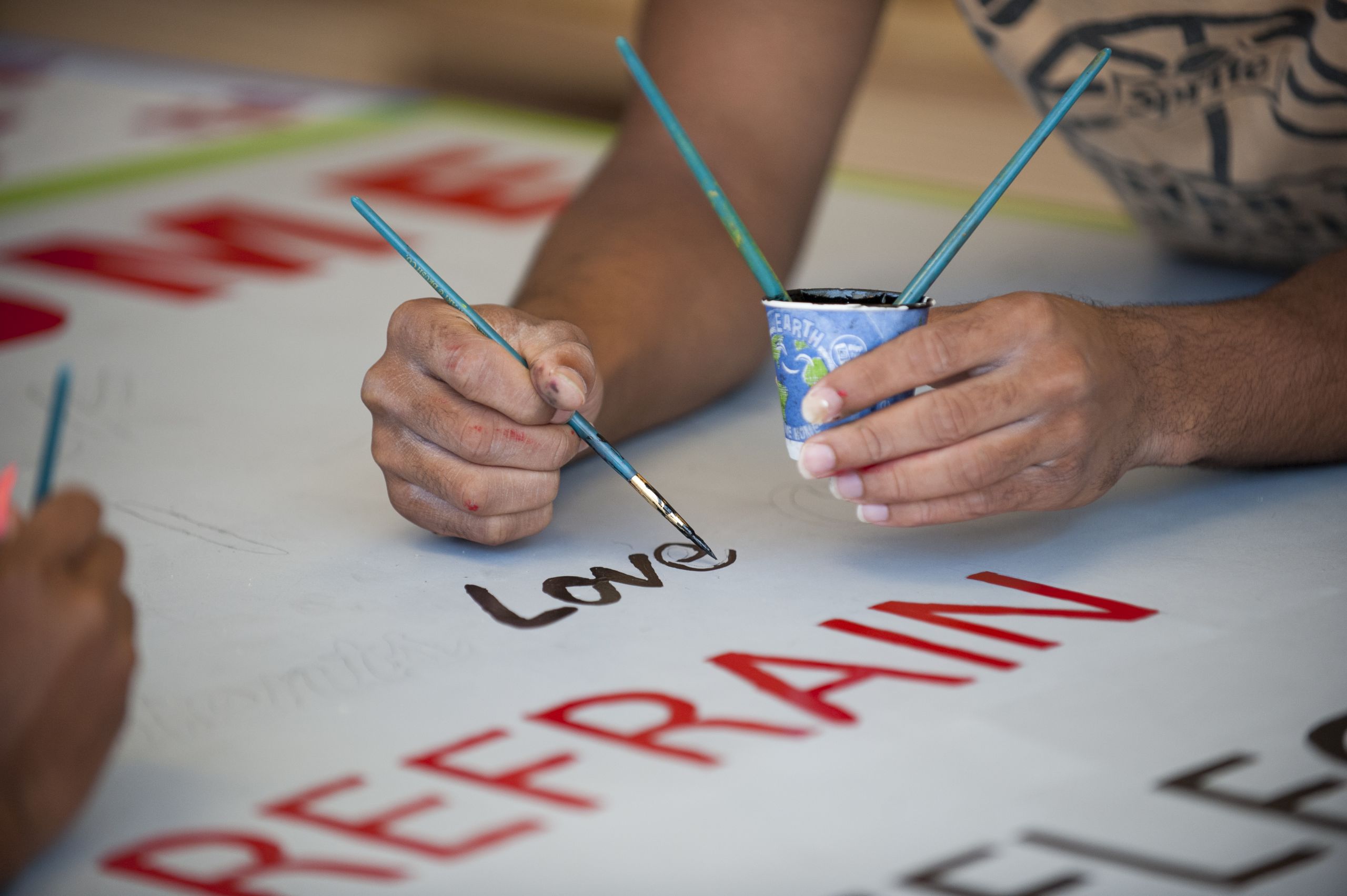 One hand holds a small paper cup of black paint, while the other paints the word "Love" on to a sign.
