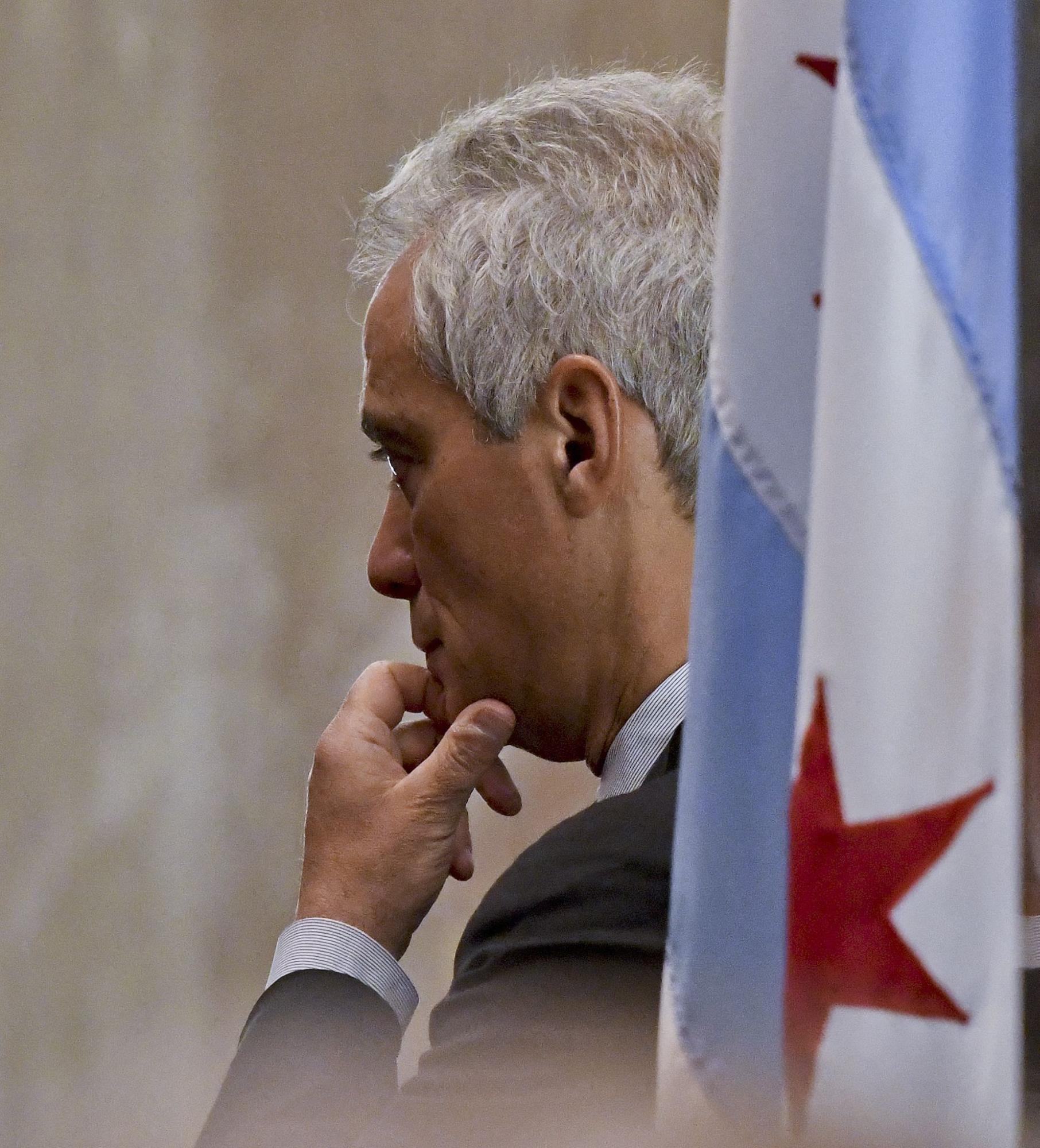 Photo of a man with grey hair in a suit from behind his left shoulder with a tan background. He holds his left hand to his chin. A Chicago flag is out of focus in the foreground.
